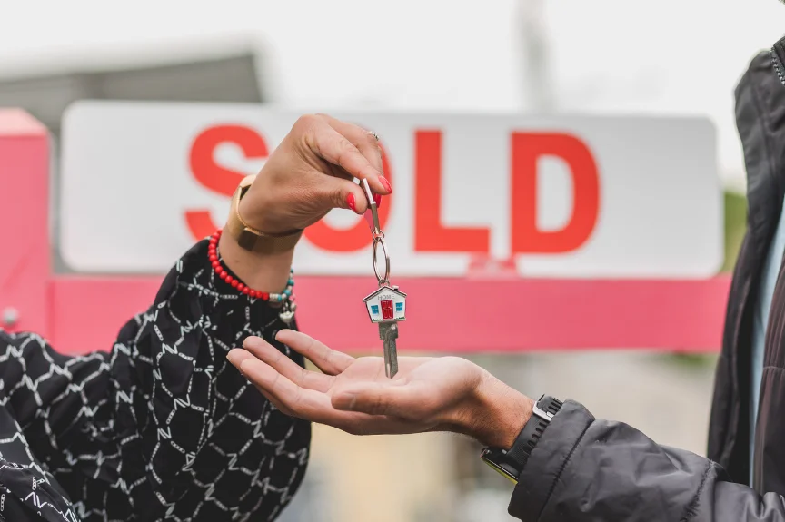 A hand passing over a house key with a SOLD sign in the background
